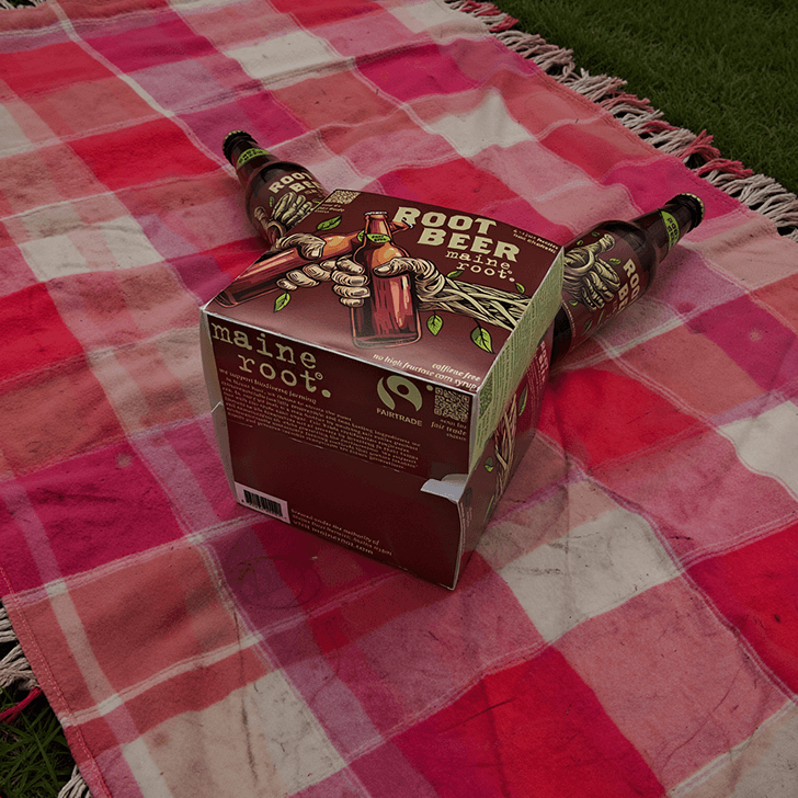 The new Maine Root packaging sits tipped over on a picnic blanket, two bottles have spilled out and the bottom of the 4-pack is visible.