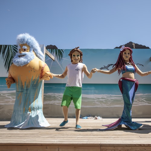 Three people in character costumes (a bearded man, a boy, and a mermaid) hold hands on a stage with a beach backdrop.