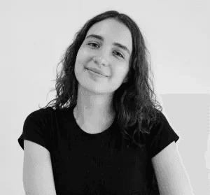Minimal studio portrait of a confident woman in a white shirt, arms crossed, on a soft gray background.