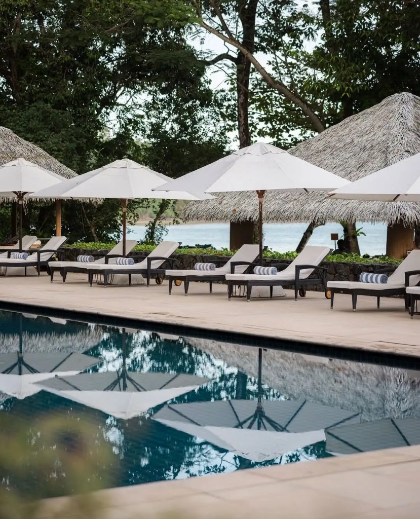 A serene pool area at The Datai Langkawi, featuring lounge chairs with umbrellas and lush greenery in the background, reflecting a commitment to a luxury experience immersed in nature.