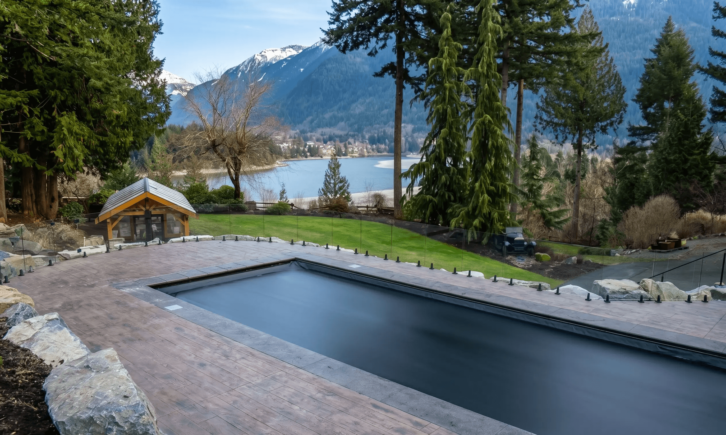 A wide-angle, bright outdoor shot of a rectangular in-ground swimming pool sealed with a smooth, matte-black automatic cover. The pool is enveloped by a light brown stamped concrete patio bordered by a frameless glass safety fence. The scenic background features a manicured green lawn, a small timber-framed shed, a wide river, and towering evergreen trees set against a dramatic backdrop of snow-capped mountains under a clear sky.