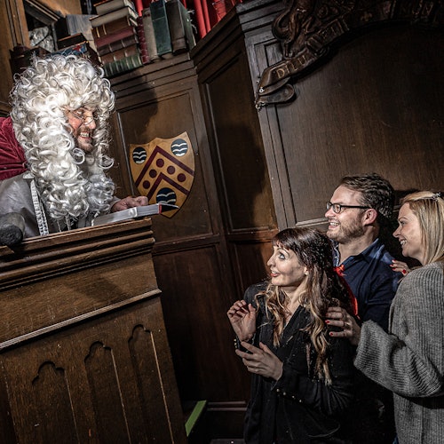 一名戴卷曲白色假发的男子站在一个高大的木制讲台上微笑,台下有三位神情愉悦的人,他们正站在一间带有深色木质墙板的房间中。