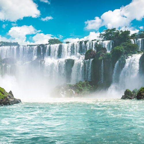 瀑布从岩石峭壁上倾泻而下，汇入宽广的河流，周围是郁郁葱葱的绿色植物，蓝天下点缀着几朵白云。