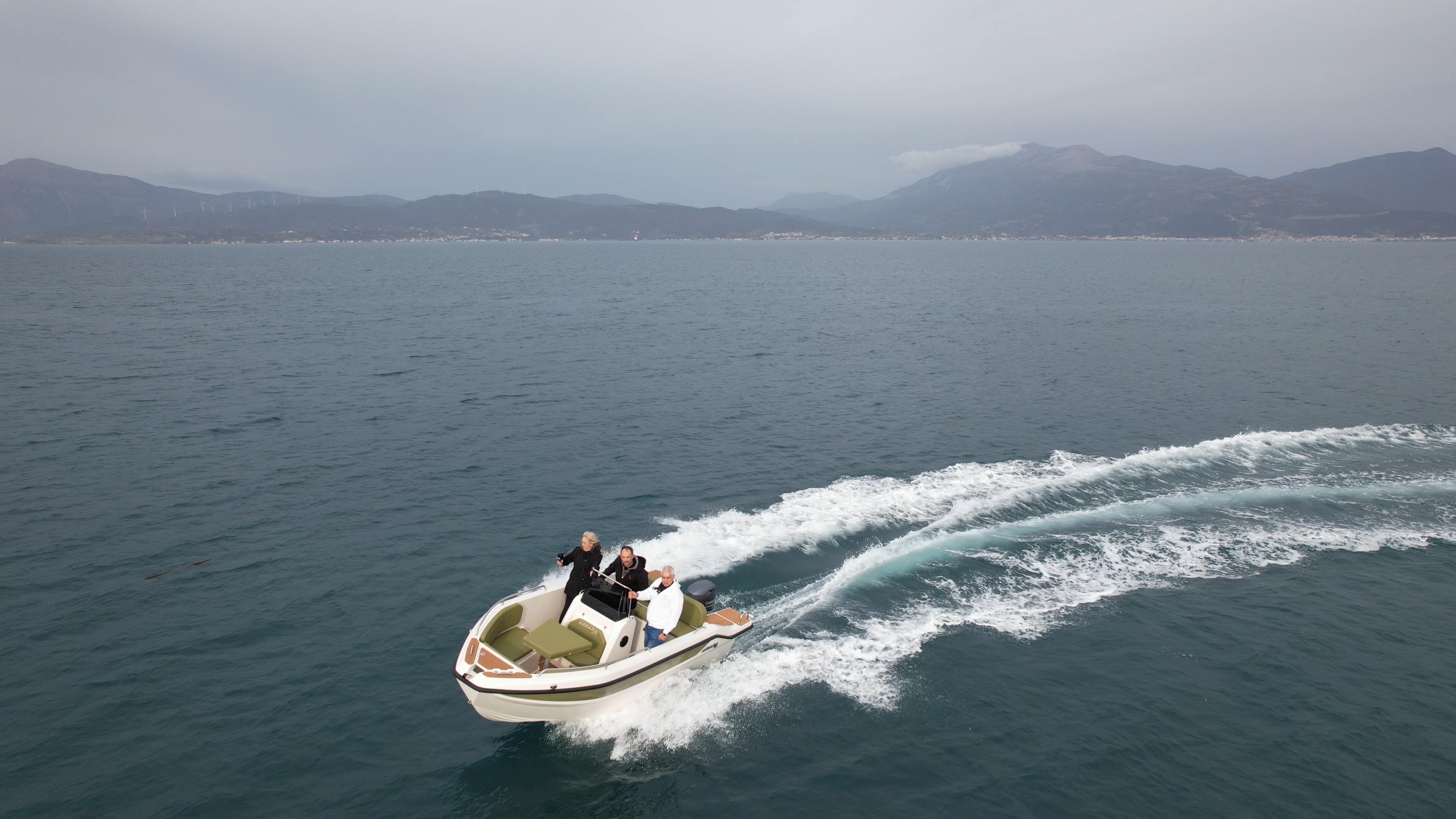 White and green speedboat with passengers cruising through Aegean waters, creating a white wake trail with mountainous Greek islands in the background.