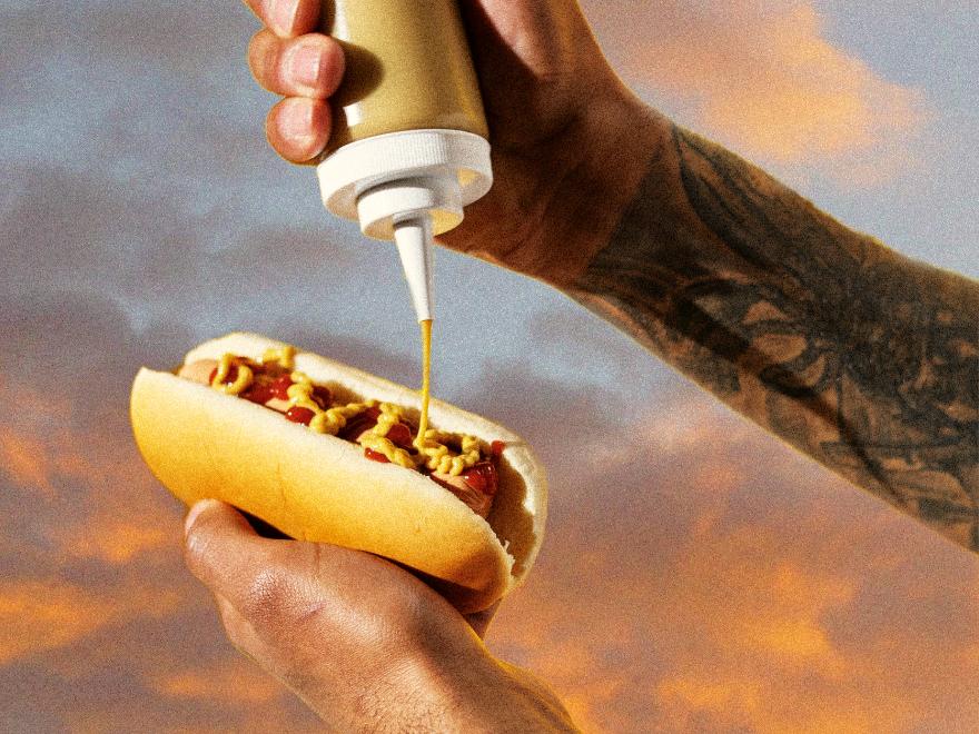 Hand pouring mustard onto hotdog with a warm sunset in the background