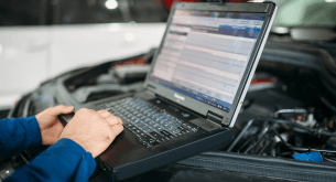 A person's hands typing on a laptop placed on top of an open car hood, with diagnostic software displayed on the screen.