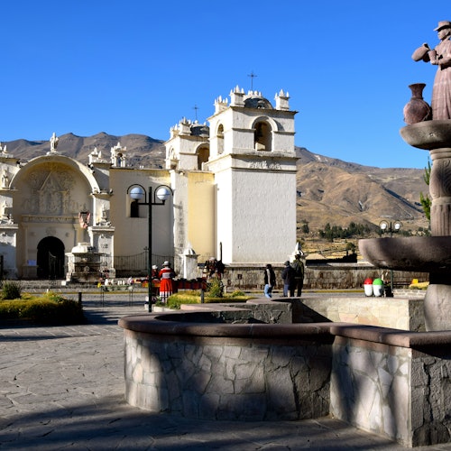 白色教堂有鐘塔，坐落在廣場上，背景是山，前景有行人和石頭噴泉。