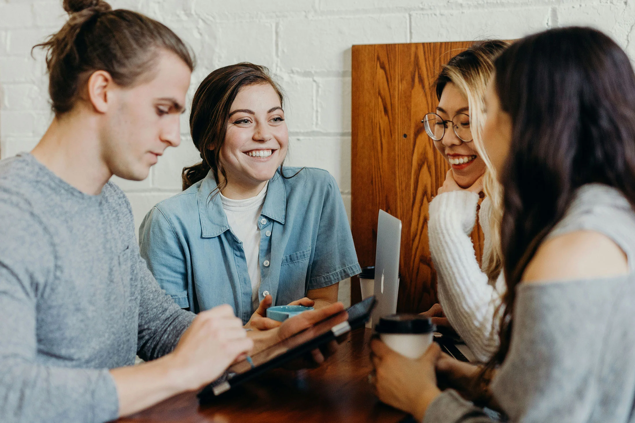 Vier junge Erwachsene sitzen in einem Café an einem Tisch, nutzen Laptop und Tablet, unterhalten sich und lachen gemeinsam, während sie an einem Projekt arbeiten.