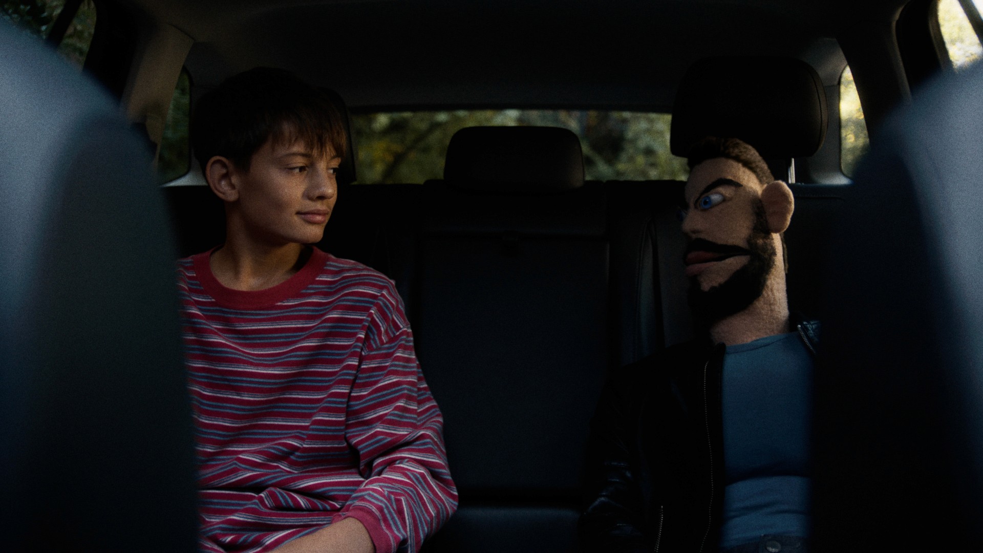 A boy in a striped shirt sits in a car's backseat, looking at a puppet with distinct eyebrows, a beard and leather jacket.