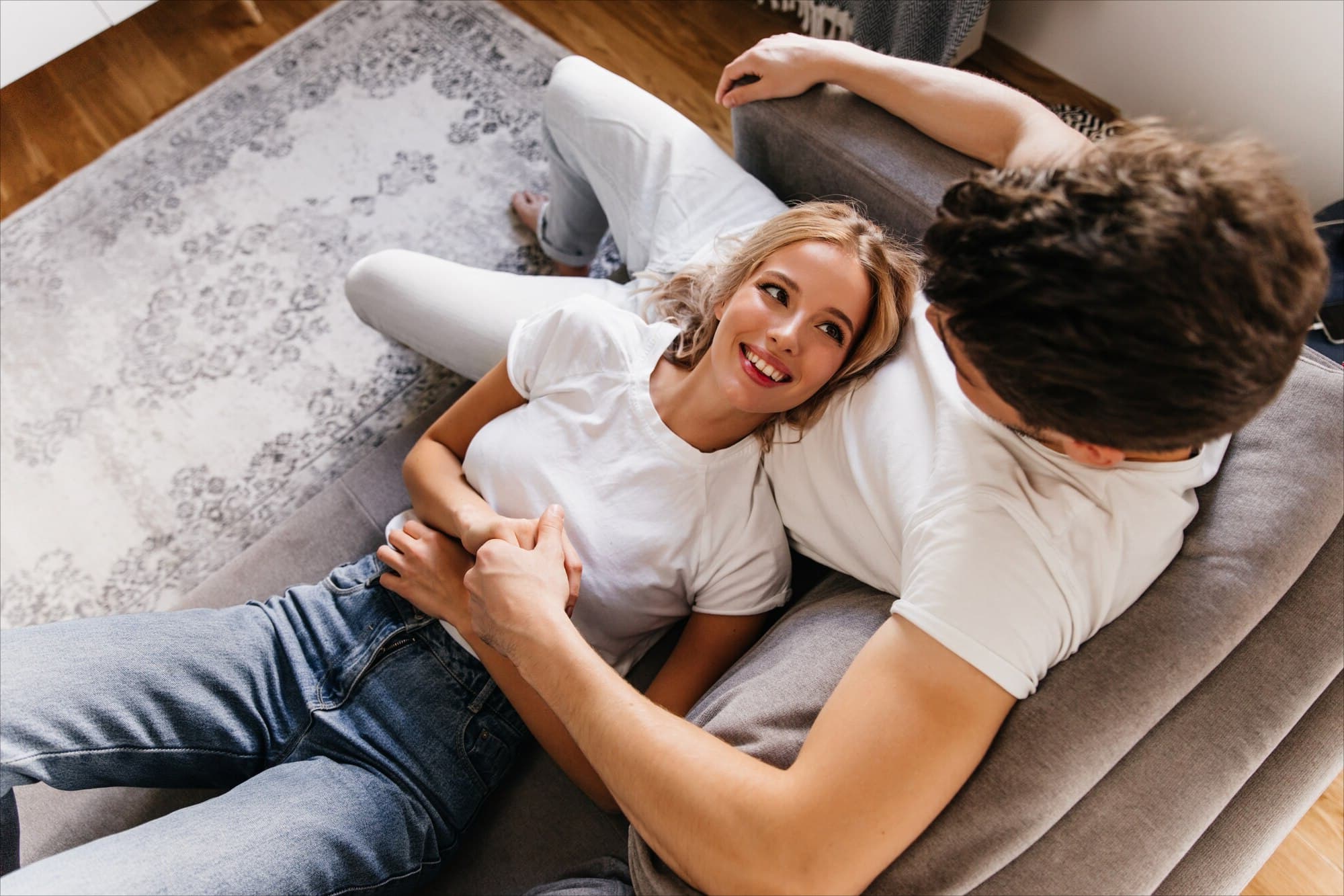 A young couple relaxes together on a couch at home, smiling and holding hands in a cozy living room. The image conveys comfort, connection, and the joy of homeownership, aligning with the message of Chris Lewis Home Loans.