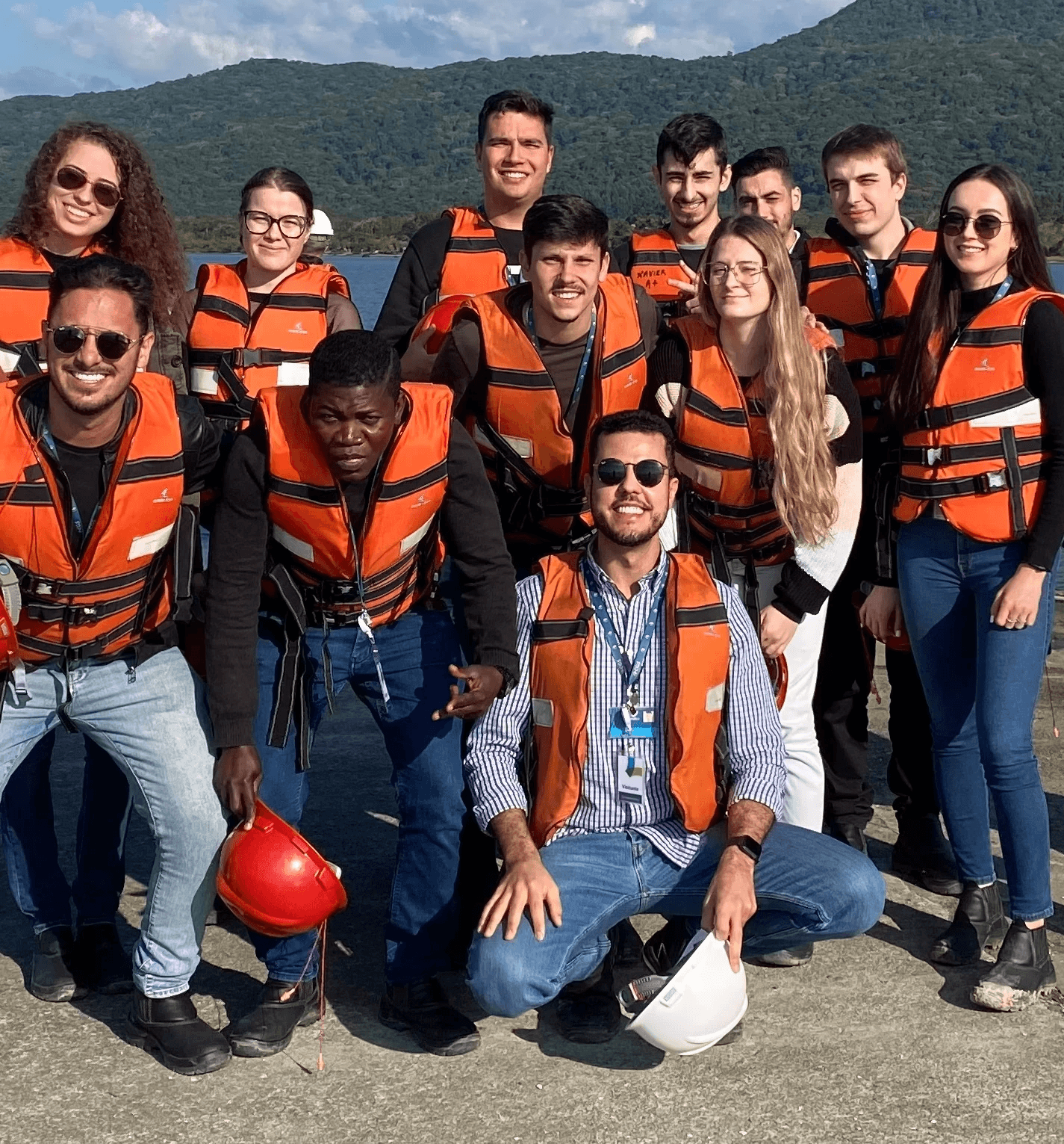 Equipe de engenheiros e técnicos sorrindo, vestindo coletes salva-vidas laranjas e segurando capacetes de proteção vermelhos e brancos durante visita técnica de campo próximo a um rio.