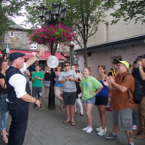 一位街头艺人在户外的路灯旁为聚集的人群表演，路灯上挂着鲜花。