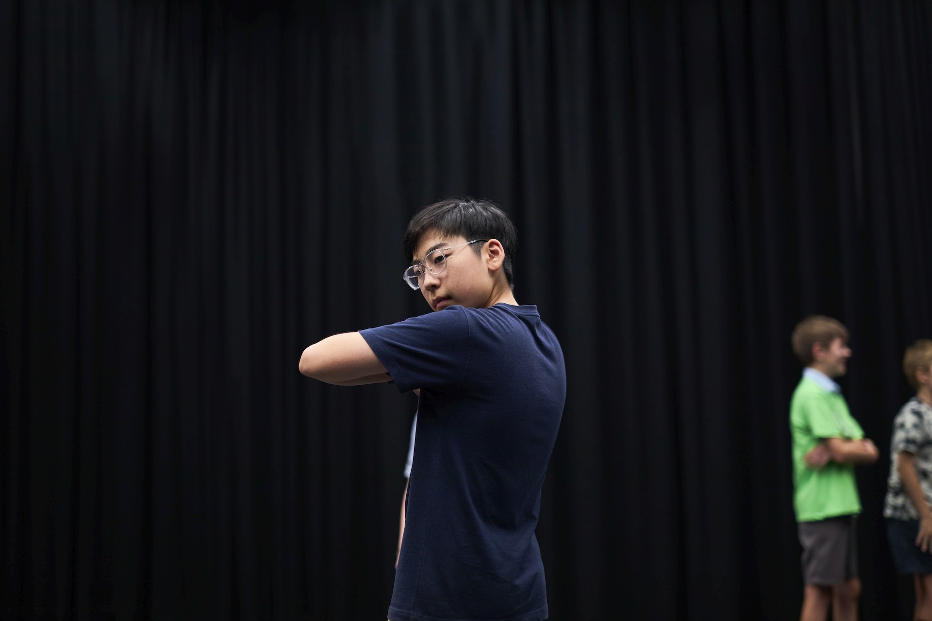 A young person in glasses looks over their shoulder while practising movement in a studio with a black curtain backdrop.