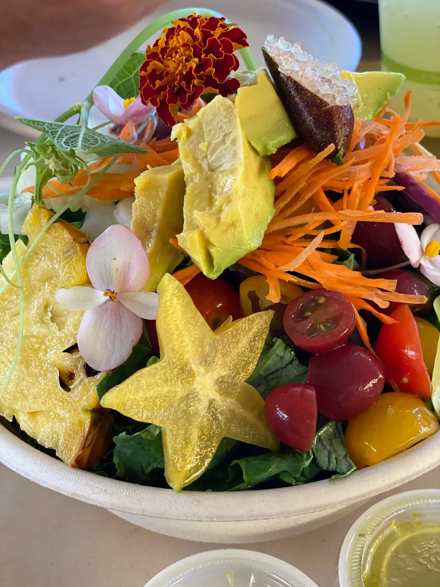 A vibrant salad bowl filled with fresh ingredients including sliced carambola, cherry tomatoes, grated carrots, avocado chunks, tropical flowers, and a honeycomb piece, all arranged artistically atop a bed of leafy greens.