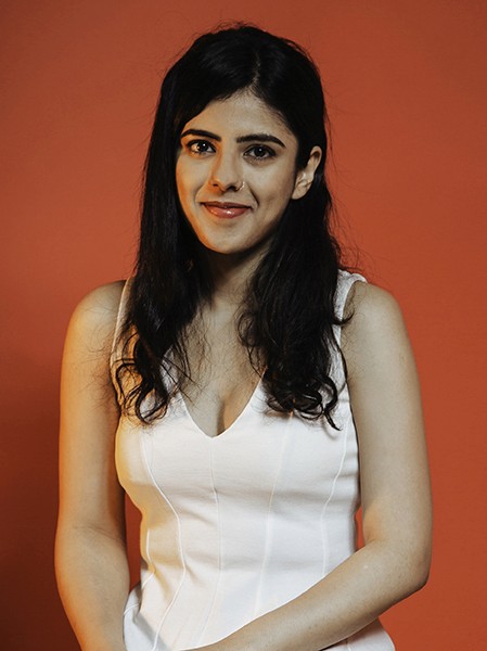 A woman with long, dark hair stands against a solid orange-brown background. She wears a sleeveless white top with a V-neck. Her gaze is directed slightly to her left, and she has a subtle smile.