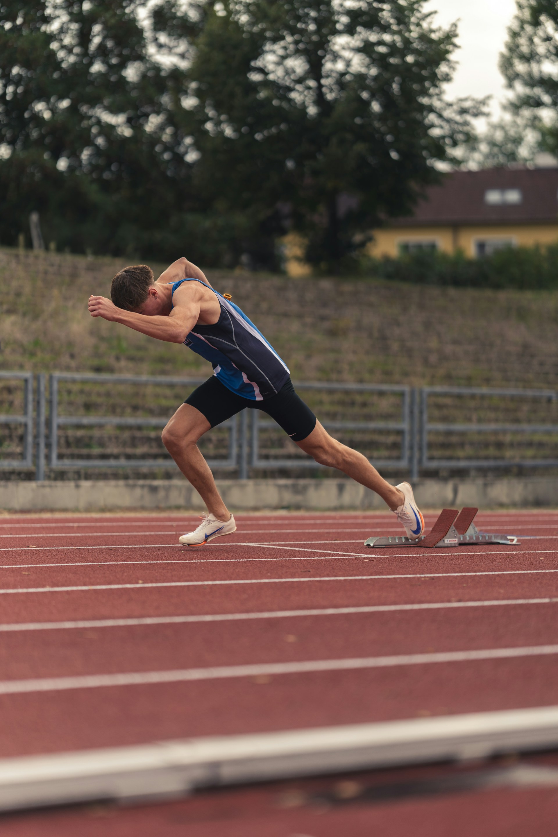 A male sprinter in a blue tank top pushing off from the starting blocks on a red running track.