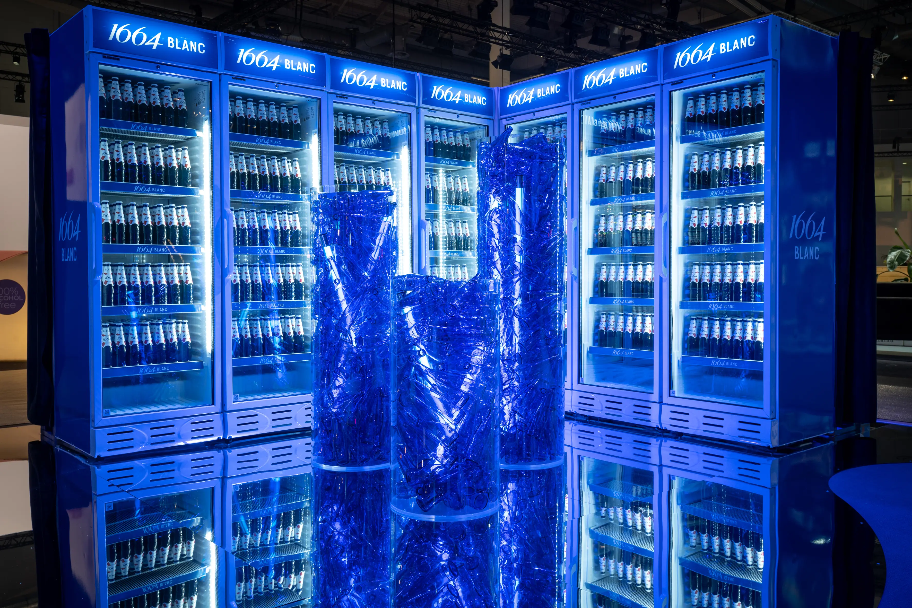 Wall of glowing 1664 Blanc fridges full of blue beer bottles behind sculptural columns of crinkled blue foil.