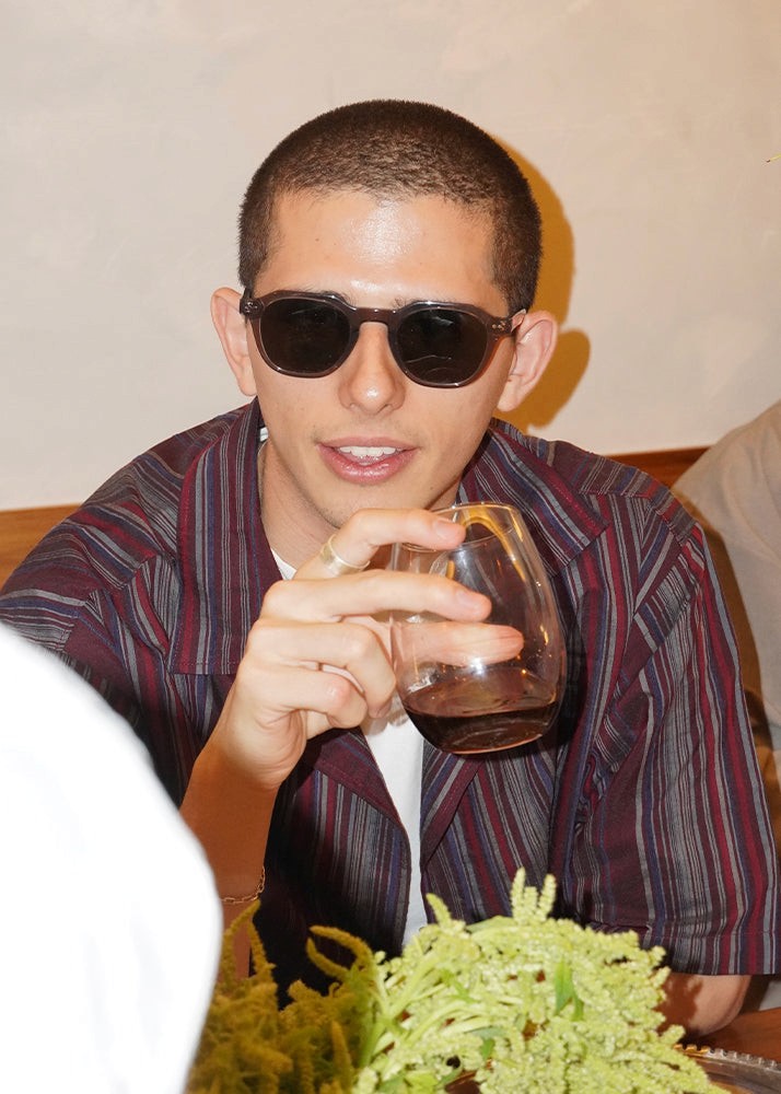 Man wearing sunglasses holding a glass of dark liquid, sitting at a table with a beige background