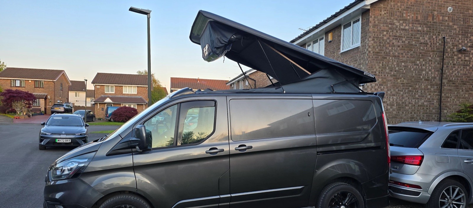 Grey Ford Transit Custom with pop-top roof raised on an evening street