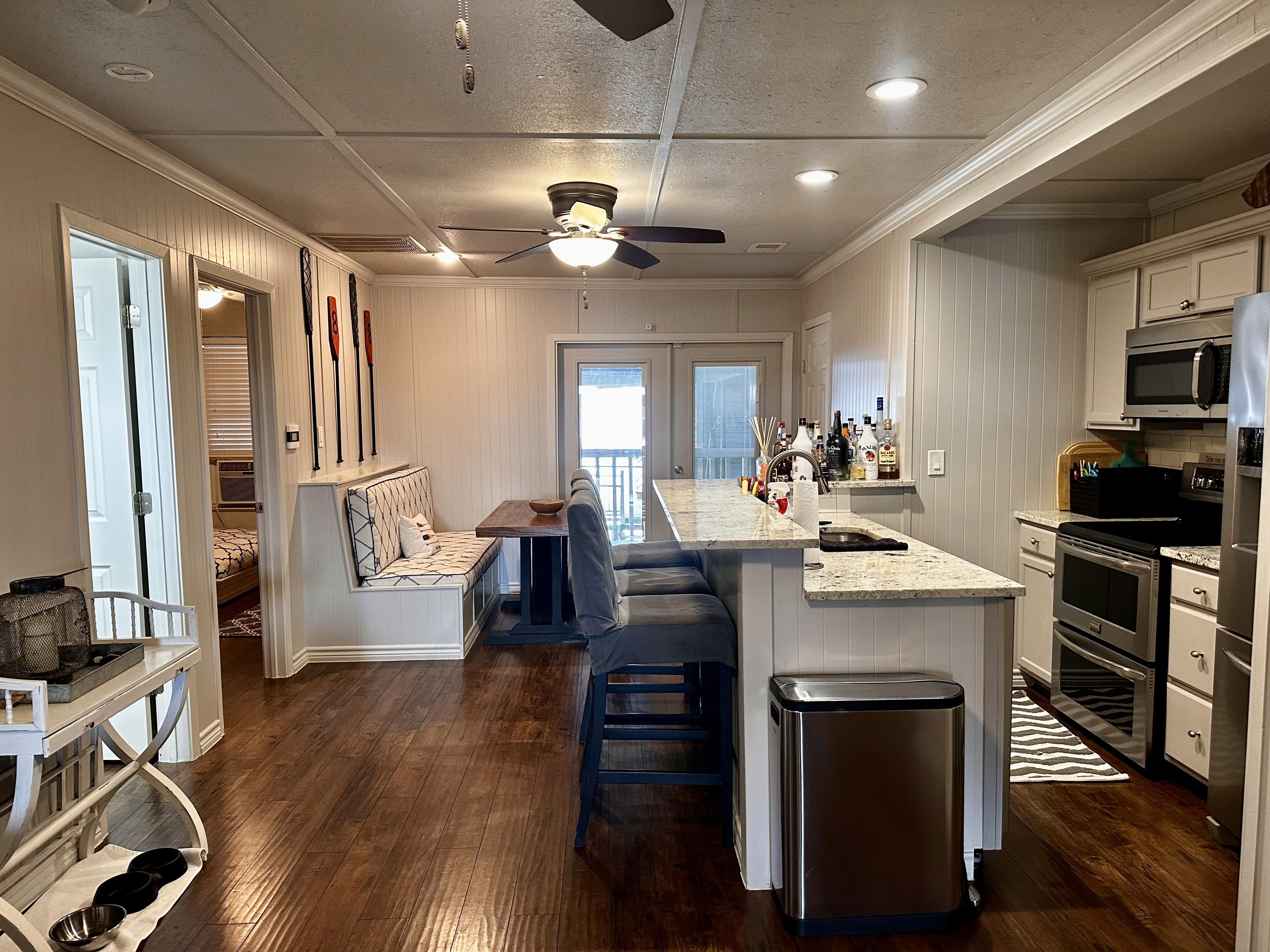 This image depicts a cozy, modern kitchen featuring a large granite kitchen island with barstool seating, stainless steel appliances, warm wood flooring, and a ceiling fan, set against a backdrop of white cabinetry and a small dining nook with bench seating.