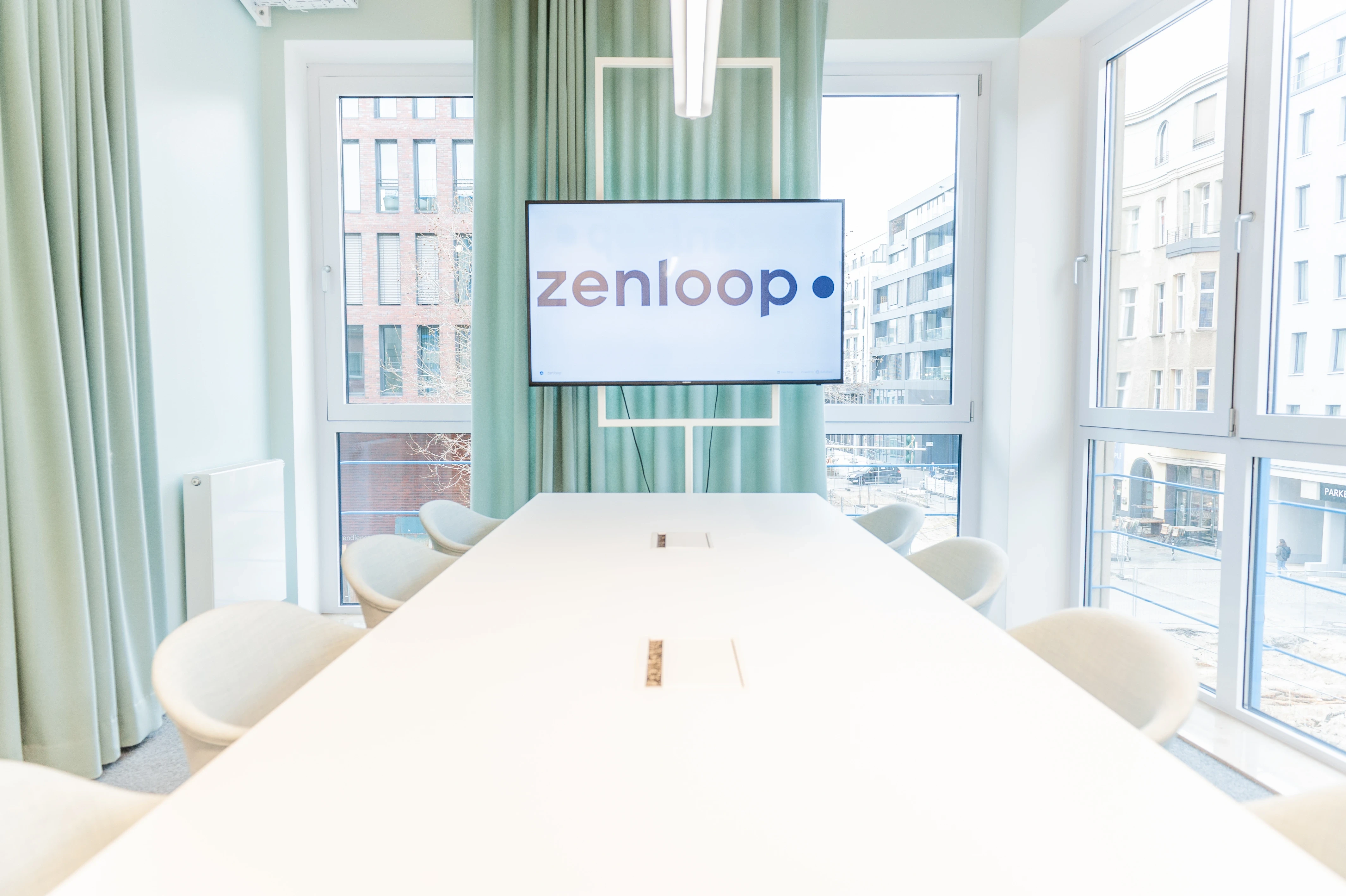 Conference room at ZENLOOP with sage-green curtains, a long white table with cream chairs and a screen showing the zenloop logo between two large windows.