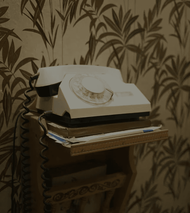 Vintage rotary telephone placed on a small wooden shelf, with papers stacked underneath and patterned wallpaper in the background.
