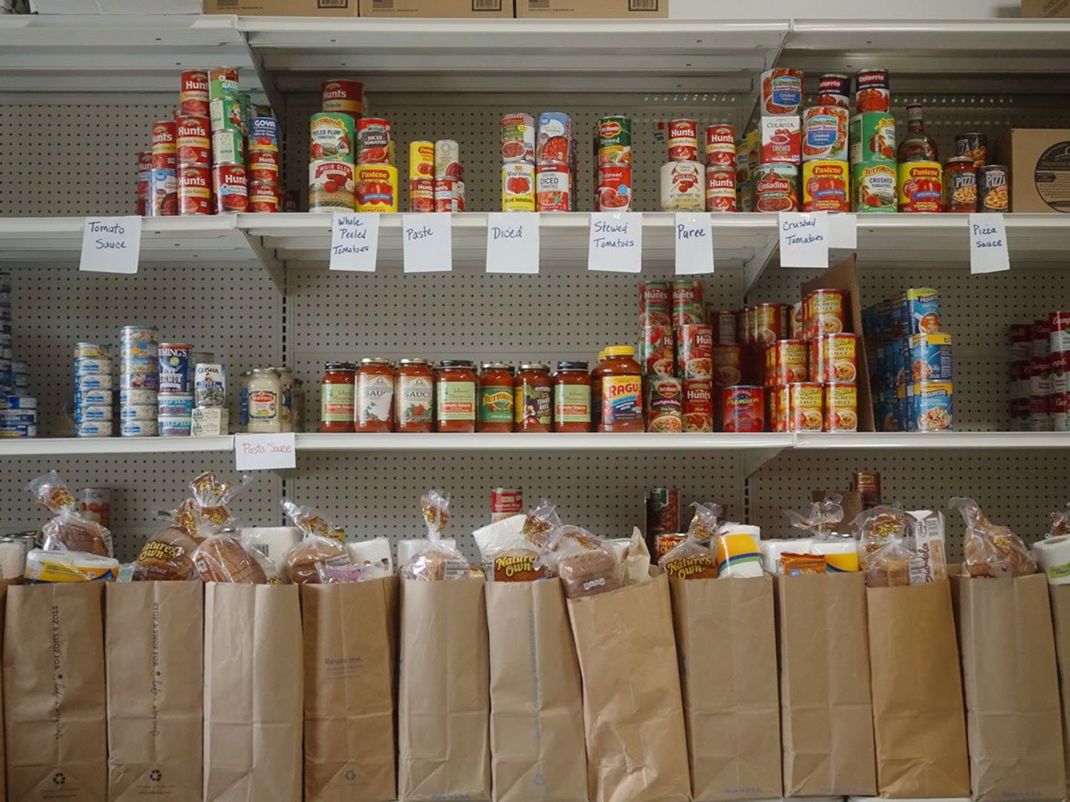 A photograph of shelves in what appears to be a food pantry or store, fully stocked with various canned tomato products and other canned goods. The shelves are labeled with signs for "Tomato Sauce," "Whole Peeled Tomatoes," "Paste," "Diced," "Stewed Tomatoes," "Puree," "Crushed Tomatoes," and "Pizza Sauce". Below the shelves, a row of brown paper bags, each containing loaves of bread, is visible.