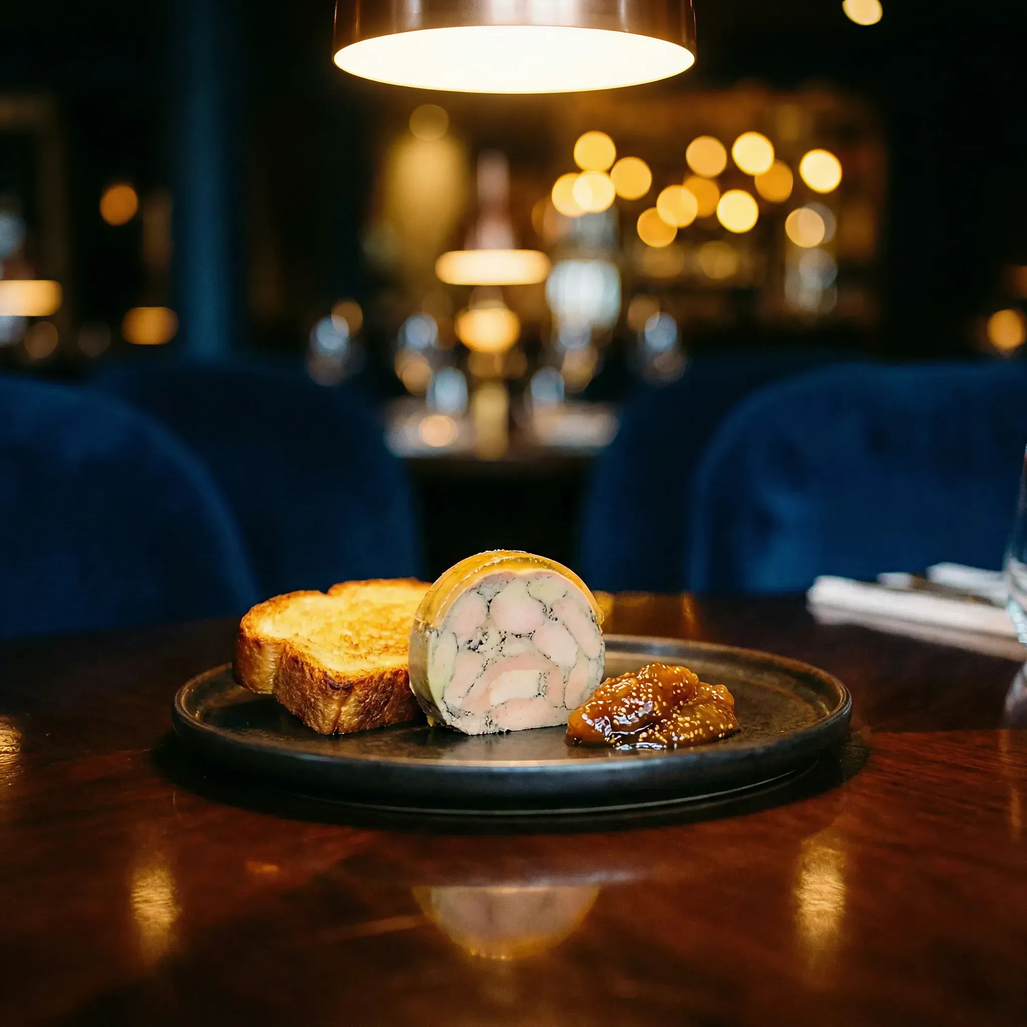 A dimly lit restaurant showcases a gourmet dish featuring toasted bread, a terrine, and chutney on a black plate, with a cozy and elegant ambiance.