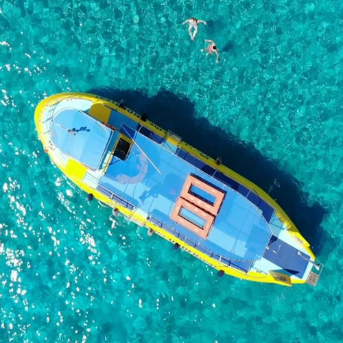 Aerial view of a large yellow and blue boat on clear turquoise water. Two swimmers are visible near the boat.