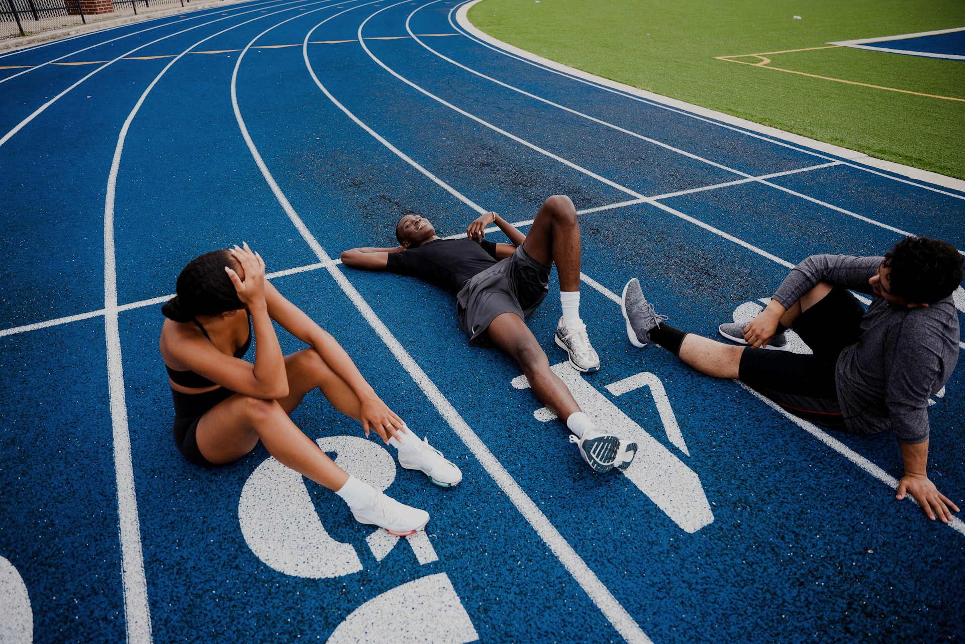 Three athletes resting on a blue running track; one lying on his back and two sitting nearby.