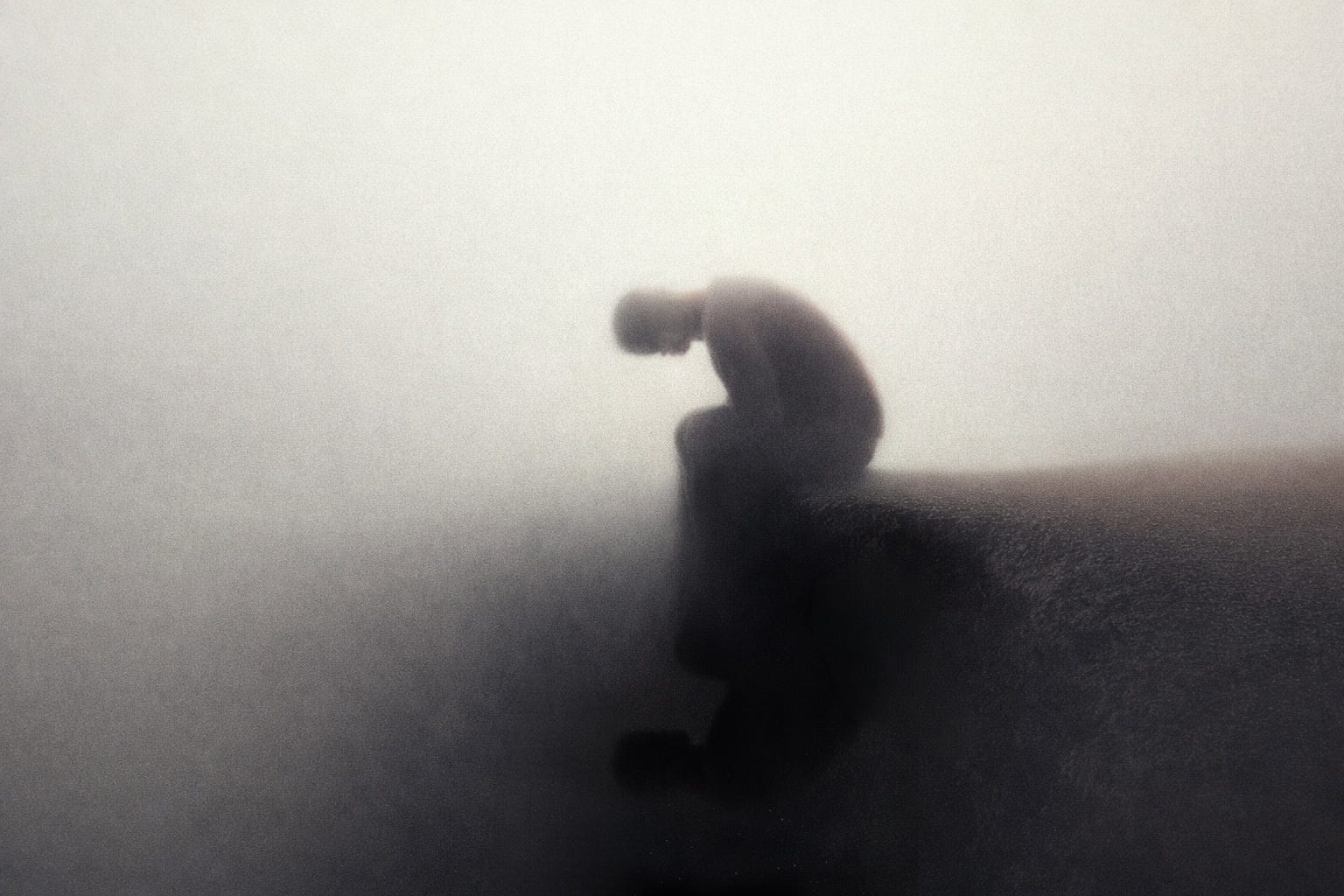 A hunched human figure enveloped in dense gray fog, gazing down at its own reflection at the bottom of the abyss.