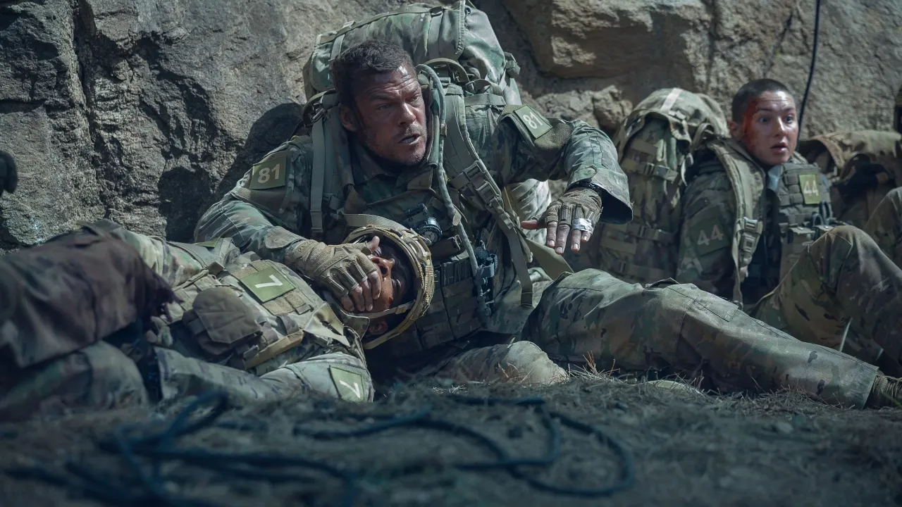 In a rugged outdoor setting, three soldiers in camouflage uniforms and tactical gear are engaged in an intense situation, with one soldier appearing concerned about a comrade lying on the ground.