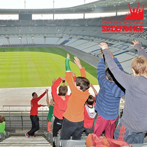 A group of children in colorful clothes raising their hands in a stadium, with a field in the background.