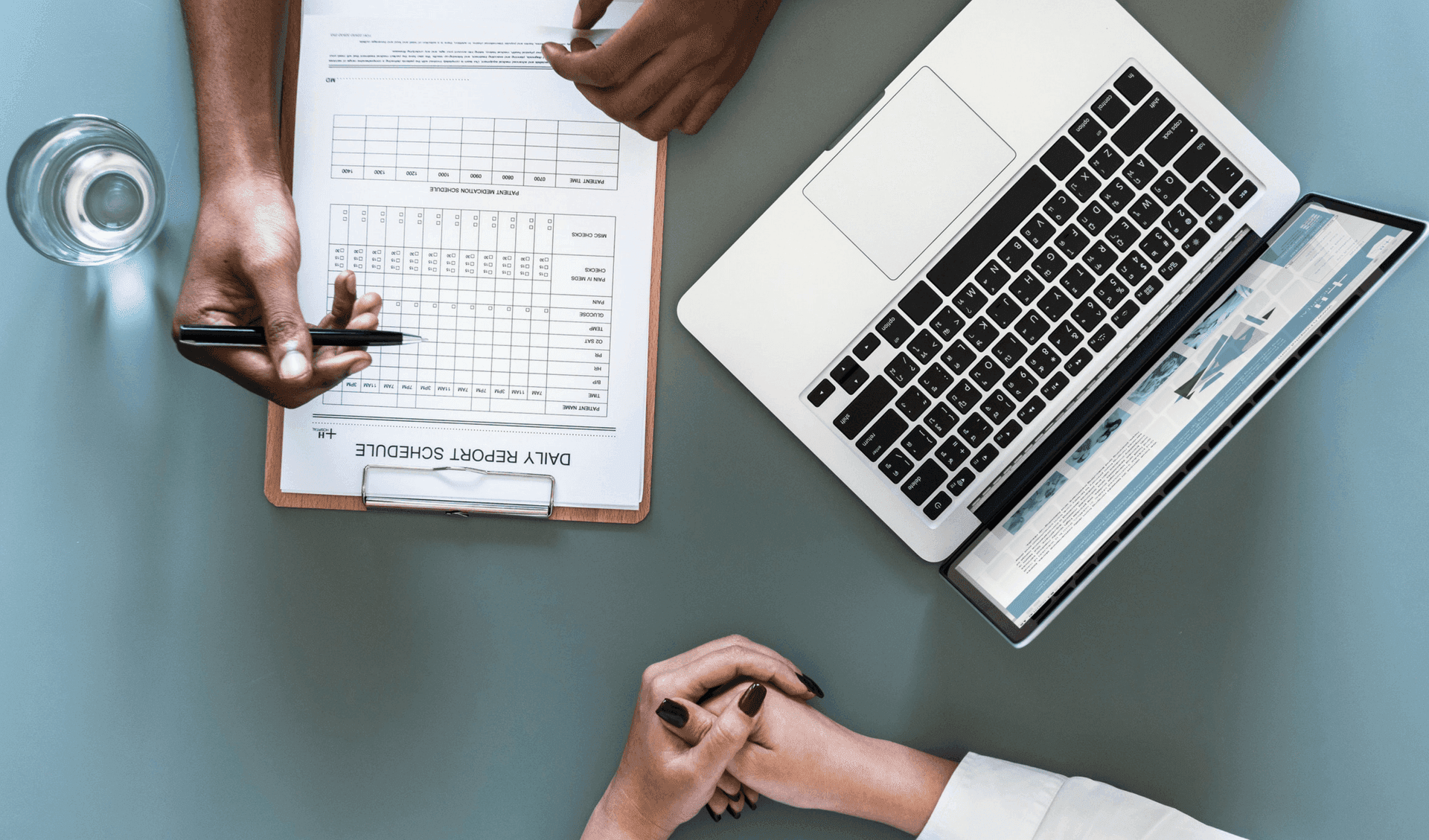 Person writing in a daily report schedule while chatting with another person with a laptop next to them