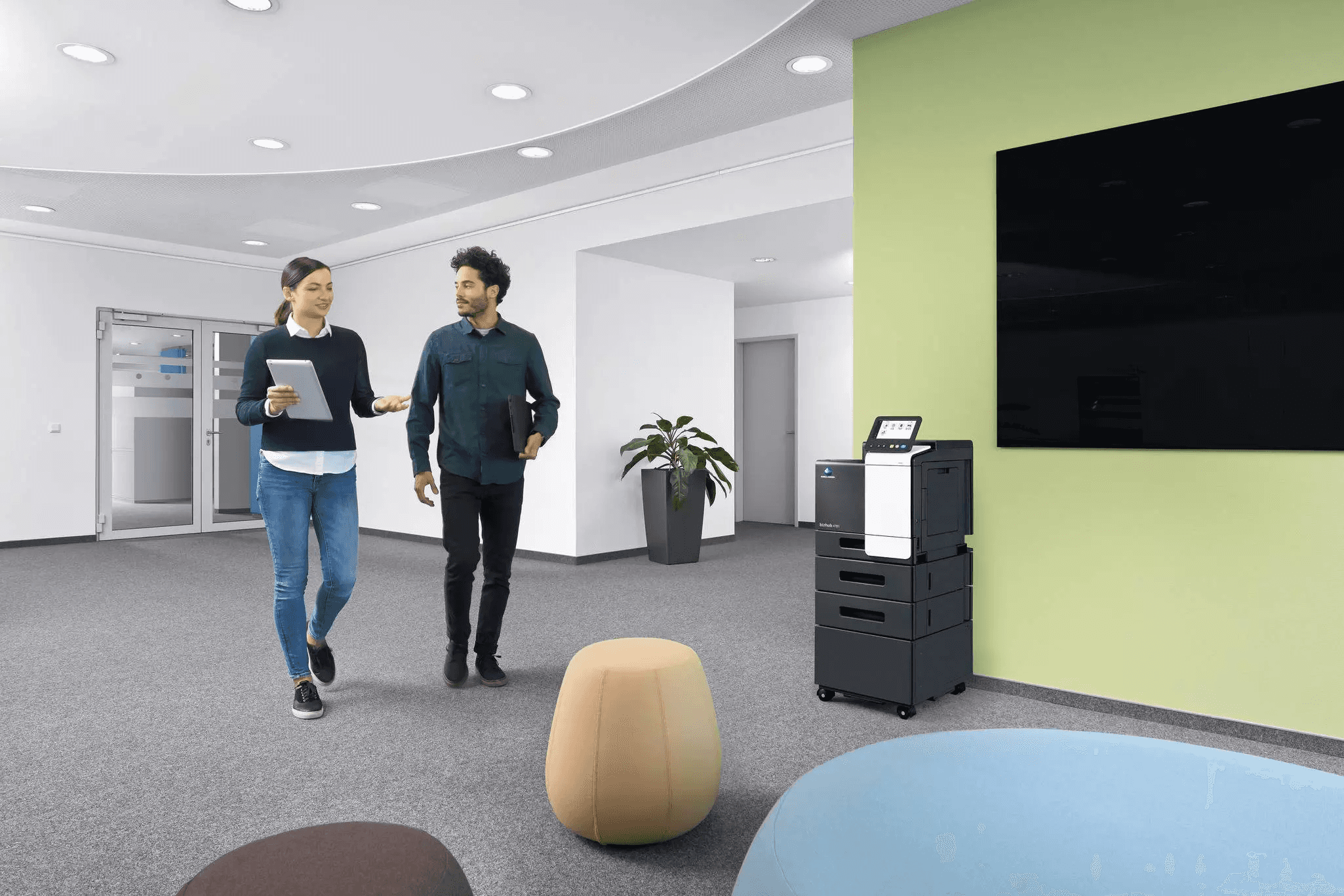 Two people walk through a modern office lobby while talking, with one holding a tablet. The space features gray carpet, a green accent wall with a large black display screen, a multifunction office printer, potted plants, glass doors, and rounded ottoman seating in the foreground.
