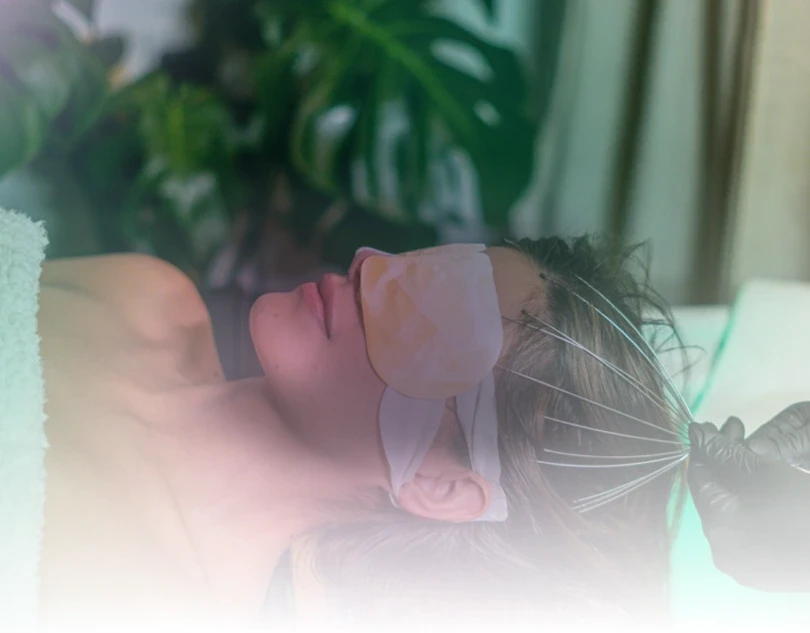 A person is lying on a spa bed with an eye mask and receiving a scalp massage, surrounded by lush green plants in a serene, relaxing environment.
