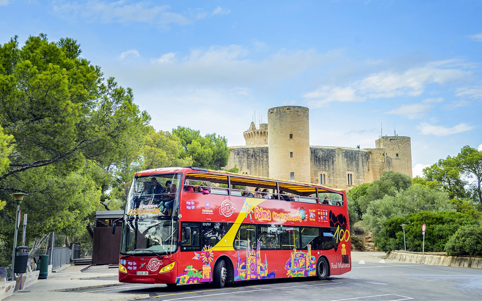 Autobús turístico HOHO frente al Castillo de Bellver, en Mallorca, rodeado de árboles.