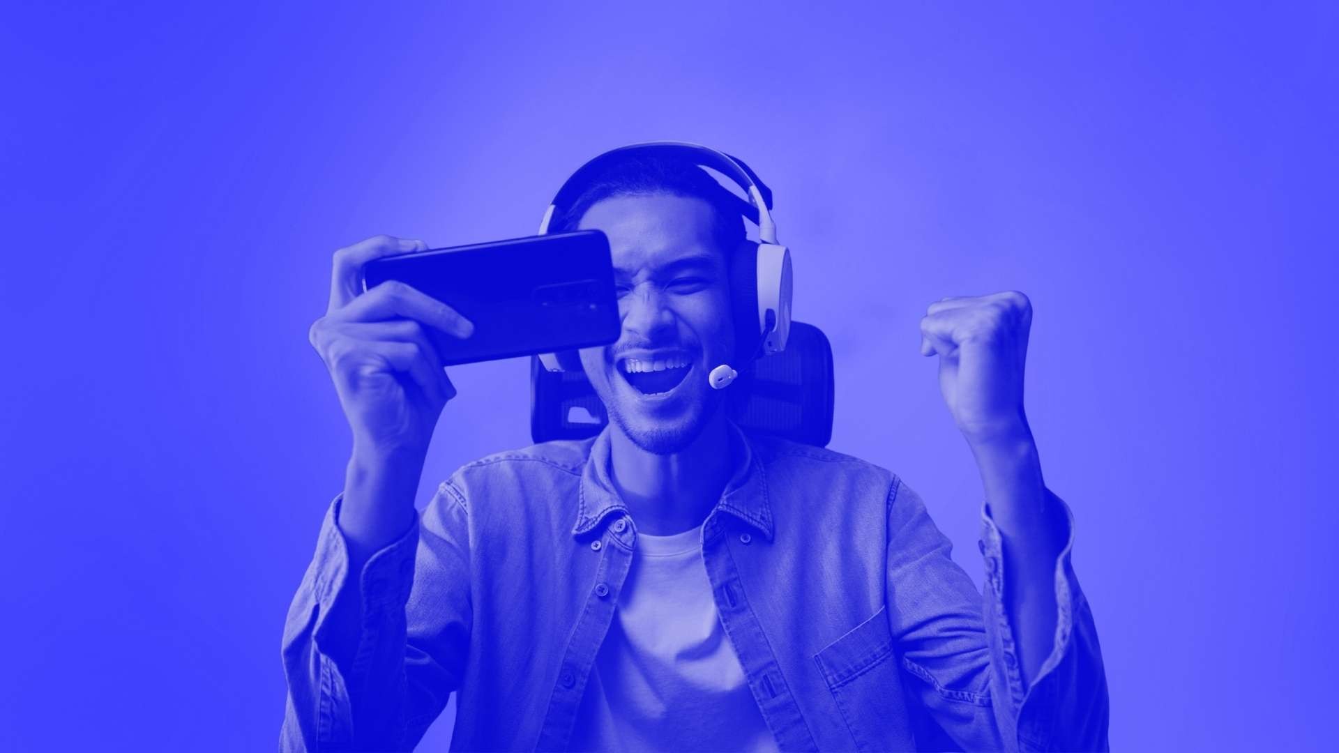 A man sitting on a chair wearing a headset and cheering at his mobile phone in his hand