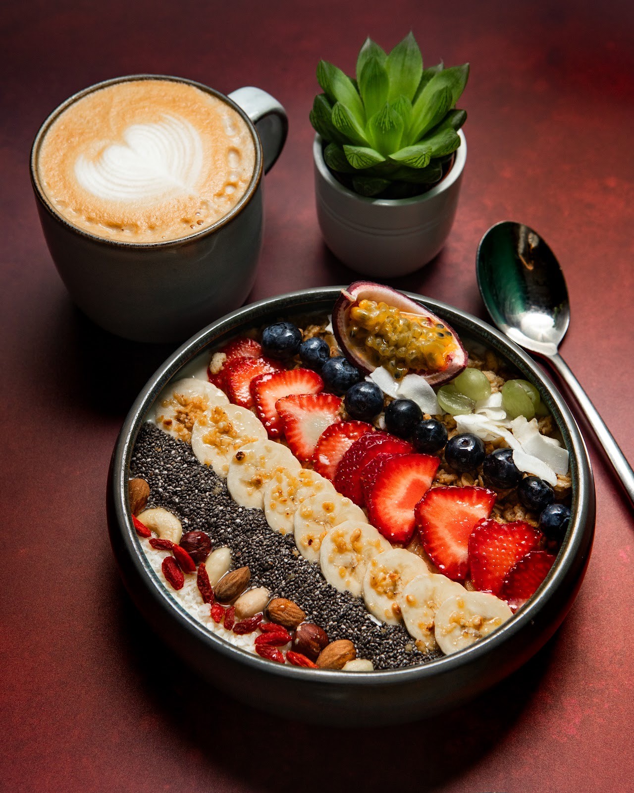 Eine farbenfrohe Acai-Bowl, belegt mit Bananenscheiben, Heidelbeeren, Erdbeeren, Himbeeren und einer grünen Frucht, daneben ein Löffel auf einem dunklen Tisch.