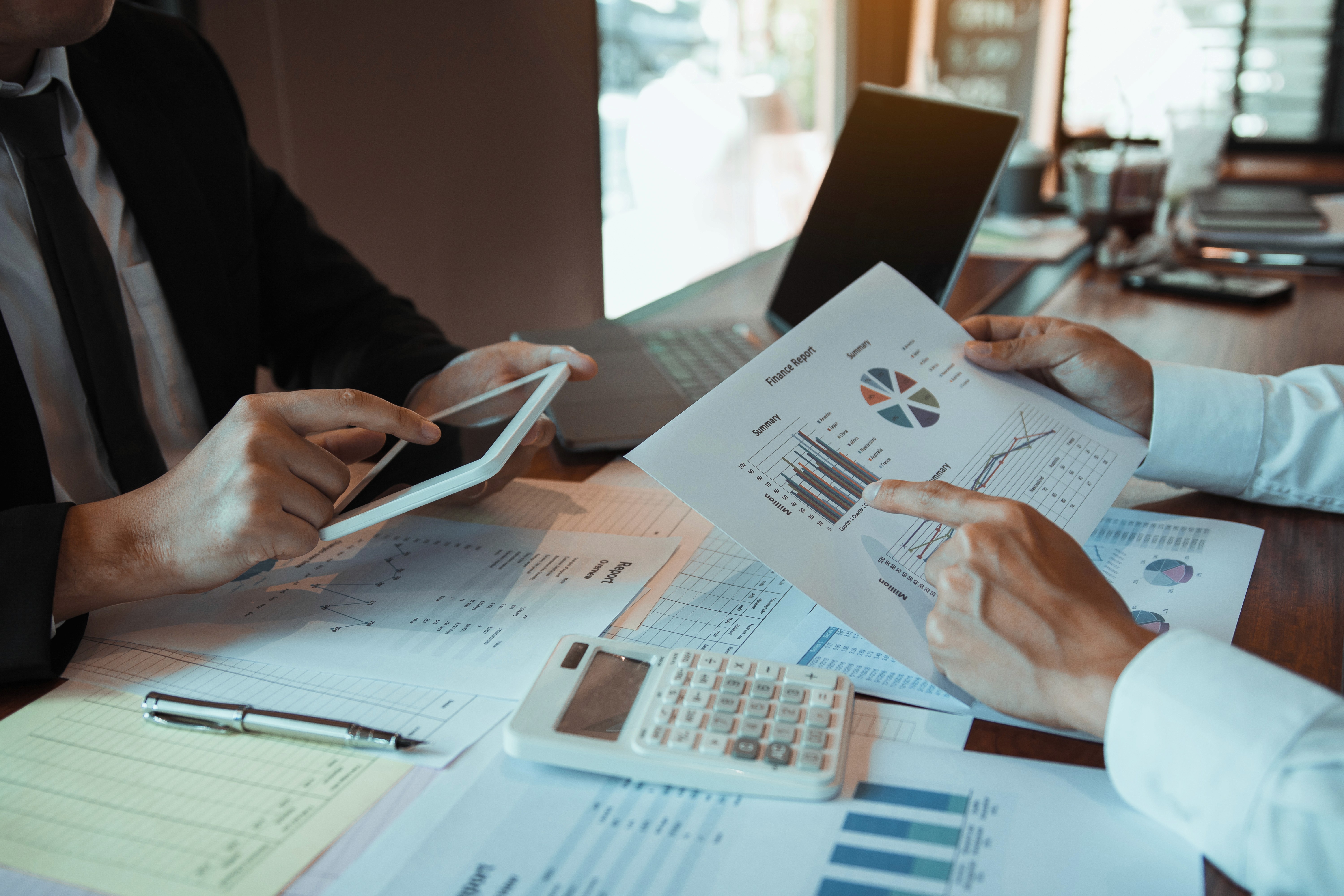 A professional tax consultant reviewing financial reports with a business owner in a modern office environment. Clean workspace, laptop displaying financial charts, documents on desk, natural daylight, premium corporate atmosphere, professional attire, trustworthy and confident expressions, high-end business photography, realistic, modern accounting firm branding.