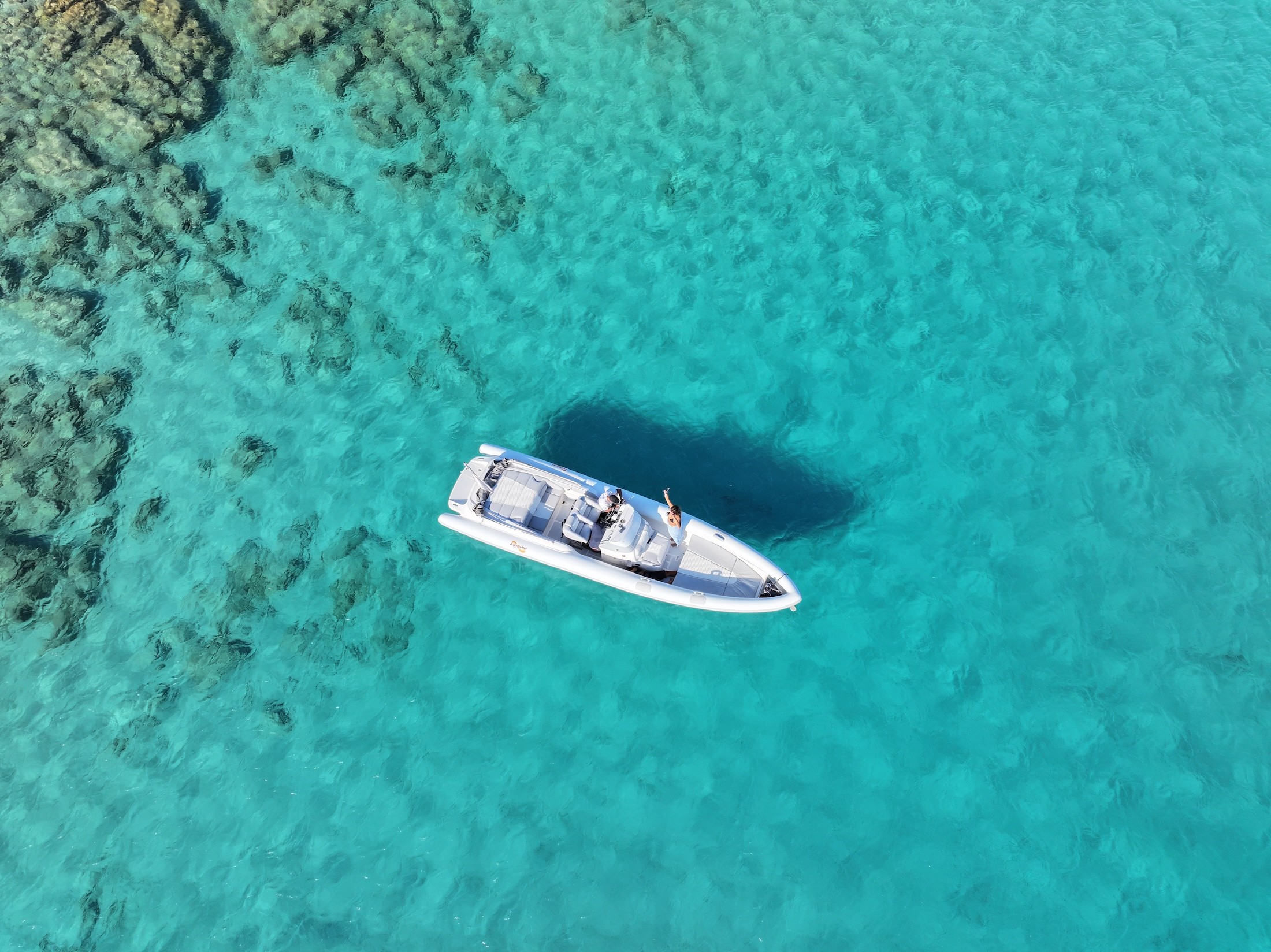 Aerial view of white Airship 30 yacht anchored in crystal-clear turquoise waters near rocky seabed formations in the Cyclades.