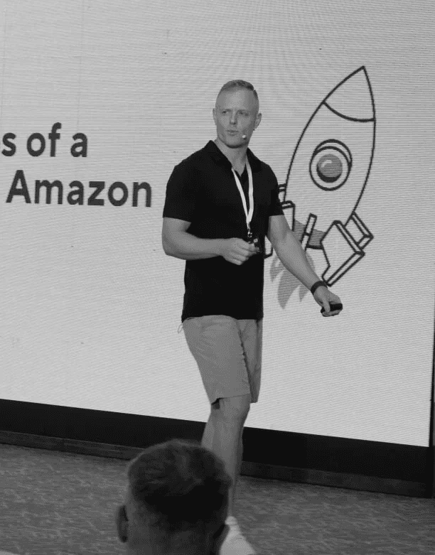 Black-and-white portrait of Riley Bennnett standing in a modern office space, smiling with hands in his pockets. He is wearing a short-sleeve button-up shirt, with blurred coworkers and hanging lights visible in the background.