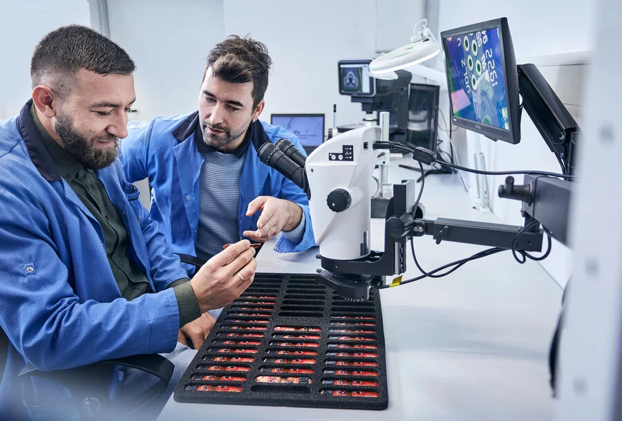 Two men in lab coats sitting at a workbench, inspecting a component. One man holds the part while the other points at it. The bench is filled with many sorted components, a microscope, and screens.