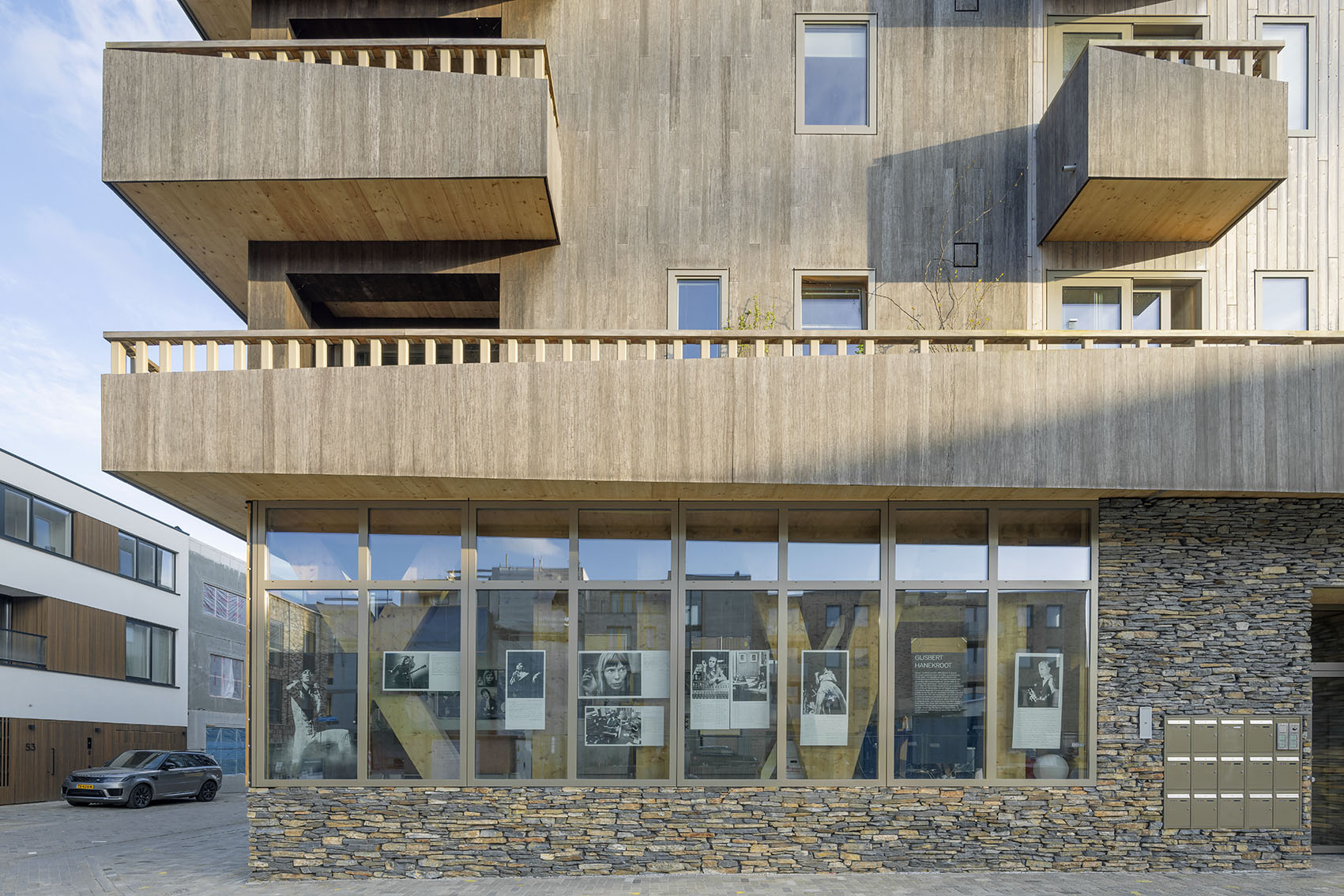Modern gebouw met houten gevel, stenen details, grote ramen en een grijze auto.
