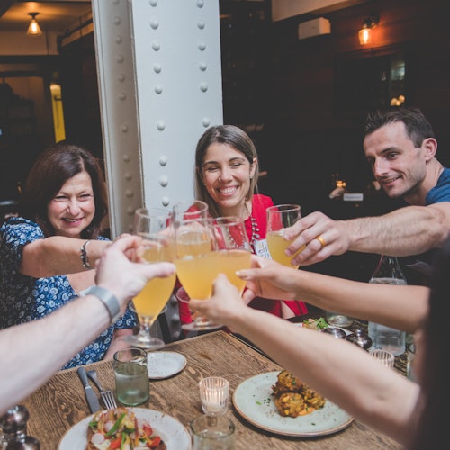 A group of people seated at a dining table clinks glasses in a toast, with plates of food and candles visible on the table.