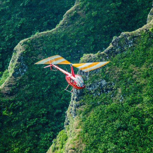 เฮลิคอปเตอร์สีแดงที่มีใบพัดลายริ้วสีเหลืองบินอยู่เหนือภูมิประเทศที่เต็มไปด้วยความเขียวขจีและเนินเขา