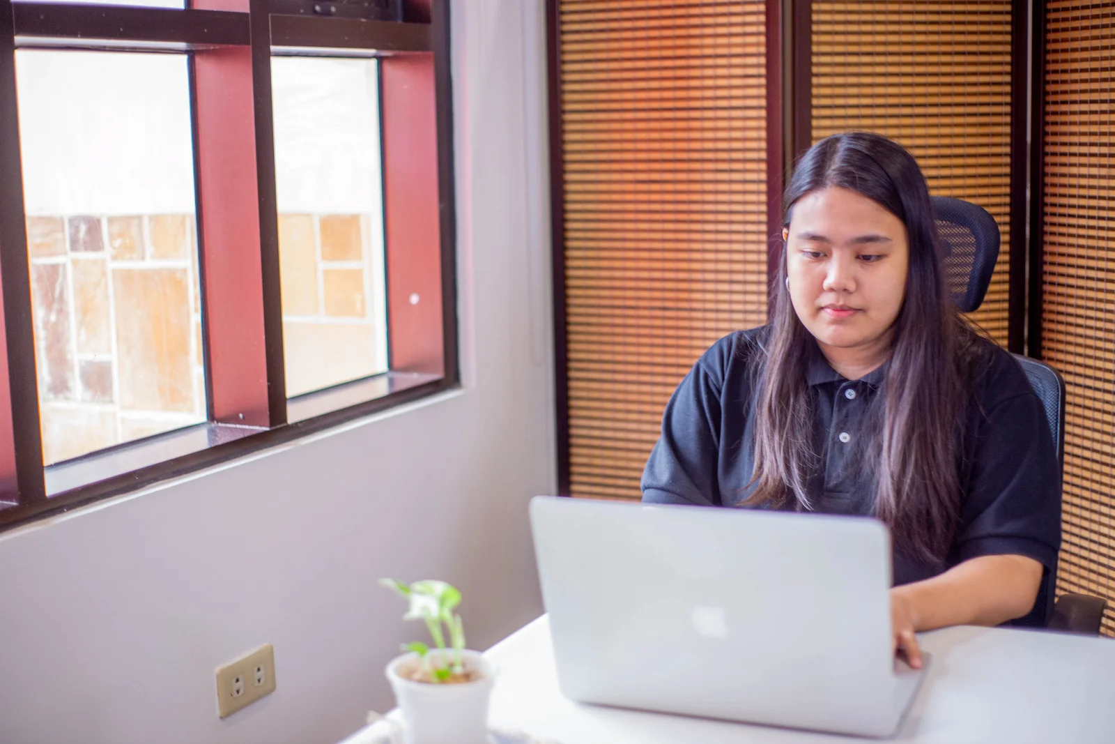 A woman sits at a desk near a window, working on a silver laptop in a bright office space.