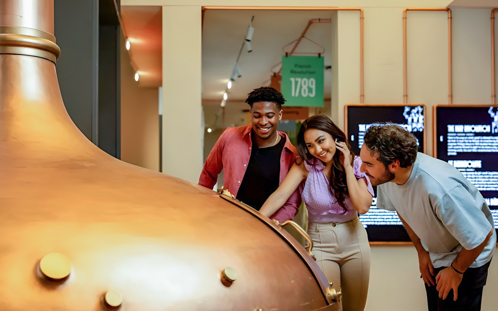 Látogatók egy nagy réz sörfőző üstöt vizsgálnak meg egy múzeumban.