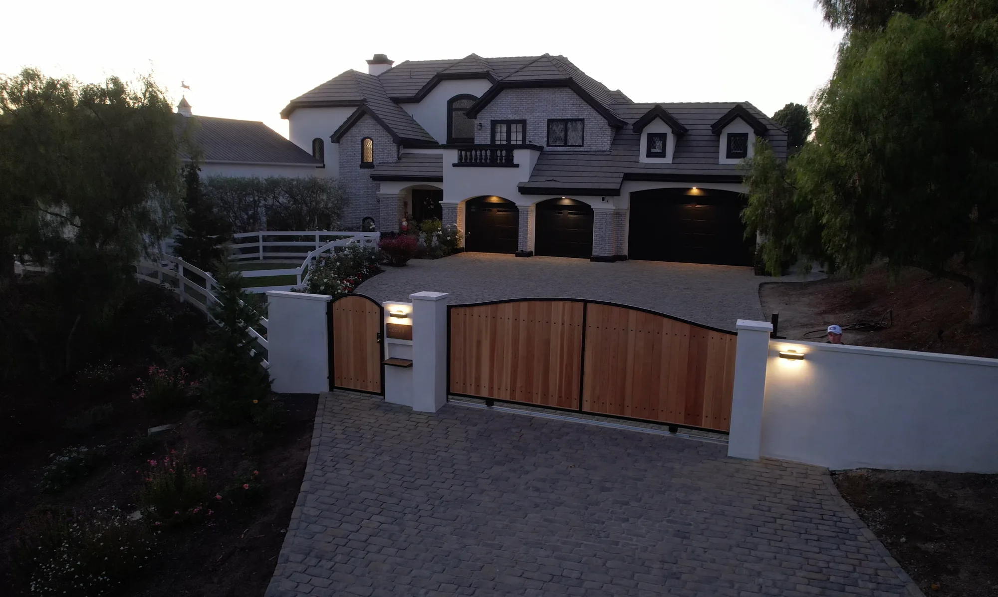 Timber entrance gates, white walls and gray paving leading toward a white home.