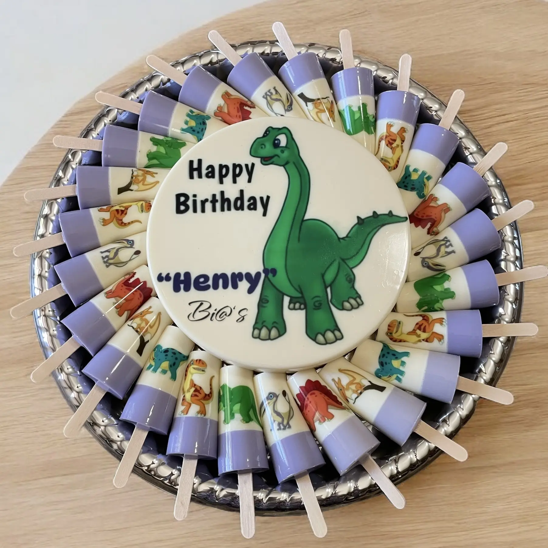 Dinosaur-themed birthday gelatin platter with a central cake displaying a friendly dinosaur and “Happy Birthday Henry” text, surrounded by matching themed gelatin cups. A fun and delightful treat for young dinosaur enthusiasts. Susa’s Sweets & Balloons.